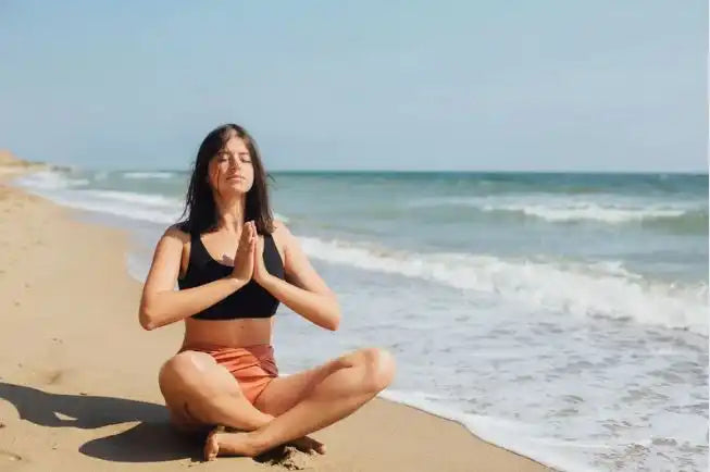 Eine Frau in einem schwarzen Sport-BH und korallenfarbenen Shorts, die auf einem Sandstrand meditieren.